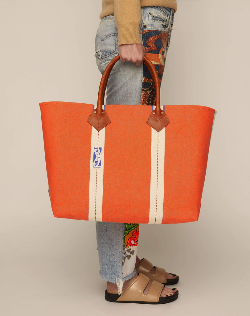 Image of person holding a classic canvas tote bag in bright orange colour with tan leather handles and contrasting natural ecru stripes.