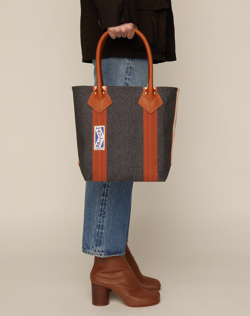 Image of person in blue jeans and leather boots holding a small classic canvas tote bag in washed black colour with tan leather handles and contrasting stripes.